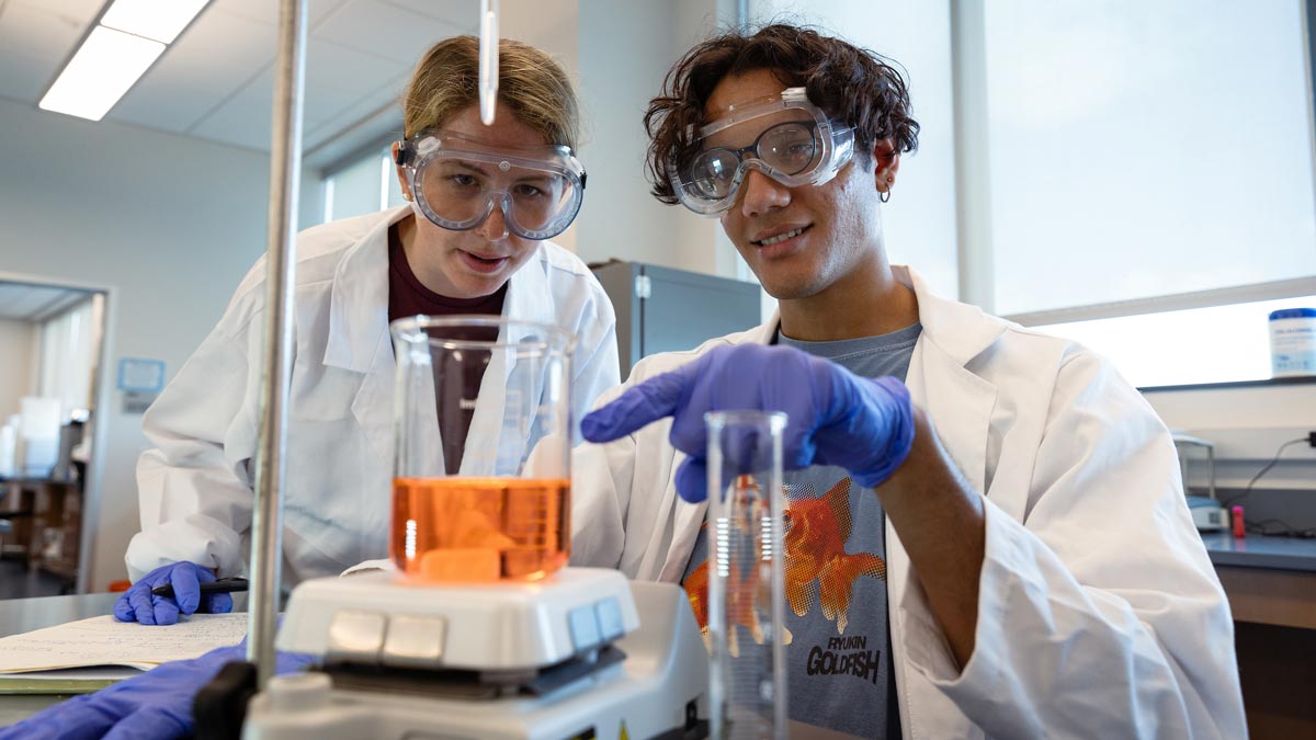 Two students in lab coats and safety goggles take a measurement of an orange liquid in a beaker.