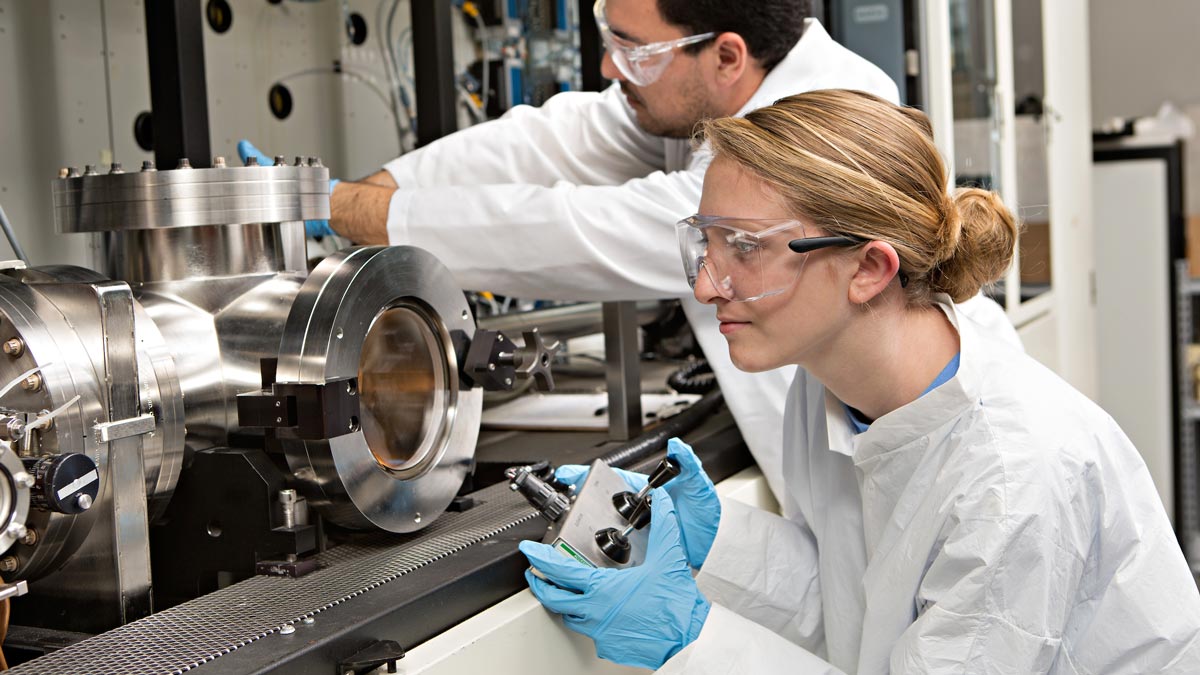 Two Aggies in lab coats and safety goggles work with industrial lab equipment.
